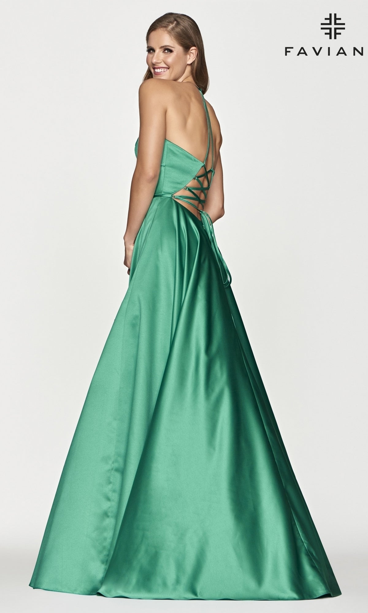 A woman stands smiling in a floor-length emerald satin Corset-Back A-Line Long Prom Dress by Faviana, seen from a rear three-quarter angle. The brand name